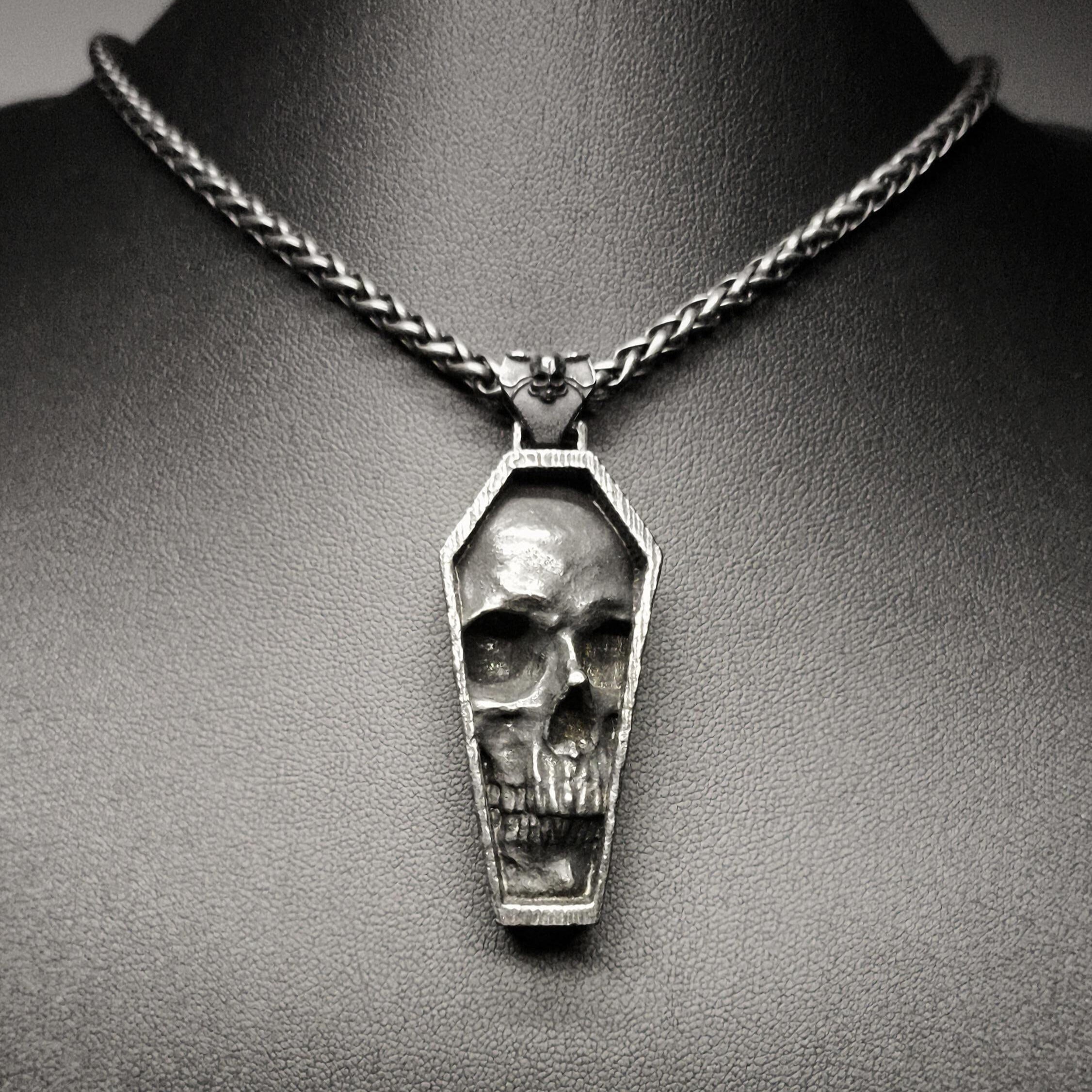 A stainless steel pendant featuring a detailed skull design in a tomb-like structure, displayed on a dark background.