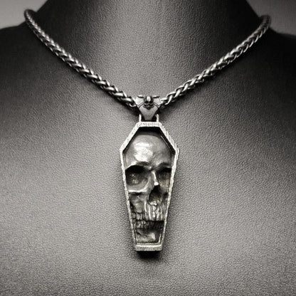 A stainless steel pendant featuring a detailed skull design in a tomb-like structure, displayed on a dark background.