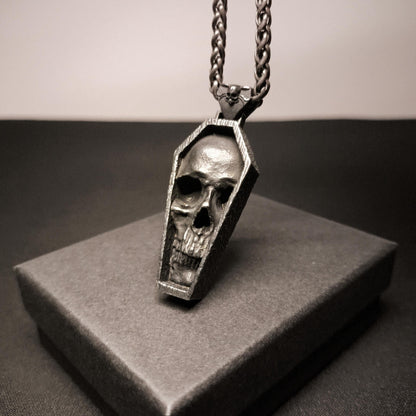 Skull-shaped pendant in a coffin design on a chain, displayed on a gray box.