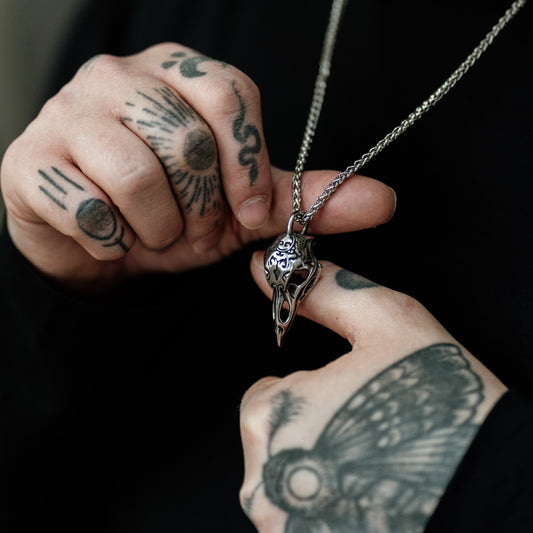 Person with tattoos holding a necklace with a bird pendant against a dark background