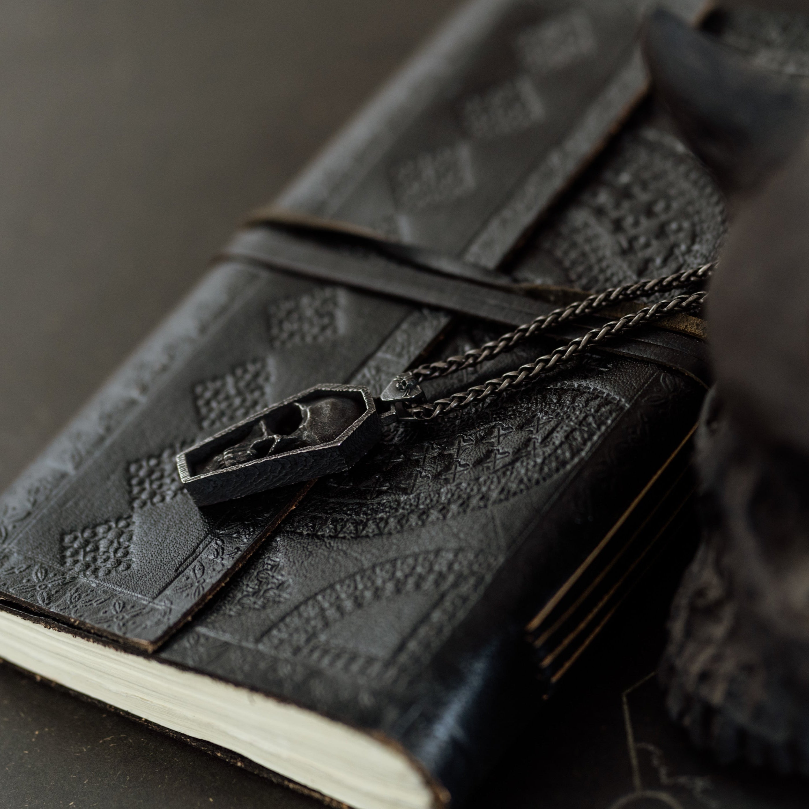 Black leather notebook with embossed design and chain, held by a dark-skinned hand on a dark surface.