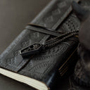 Black leather notebook with embossed design and chain, held by a dark-skinned hand on a dark surface.