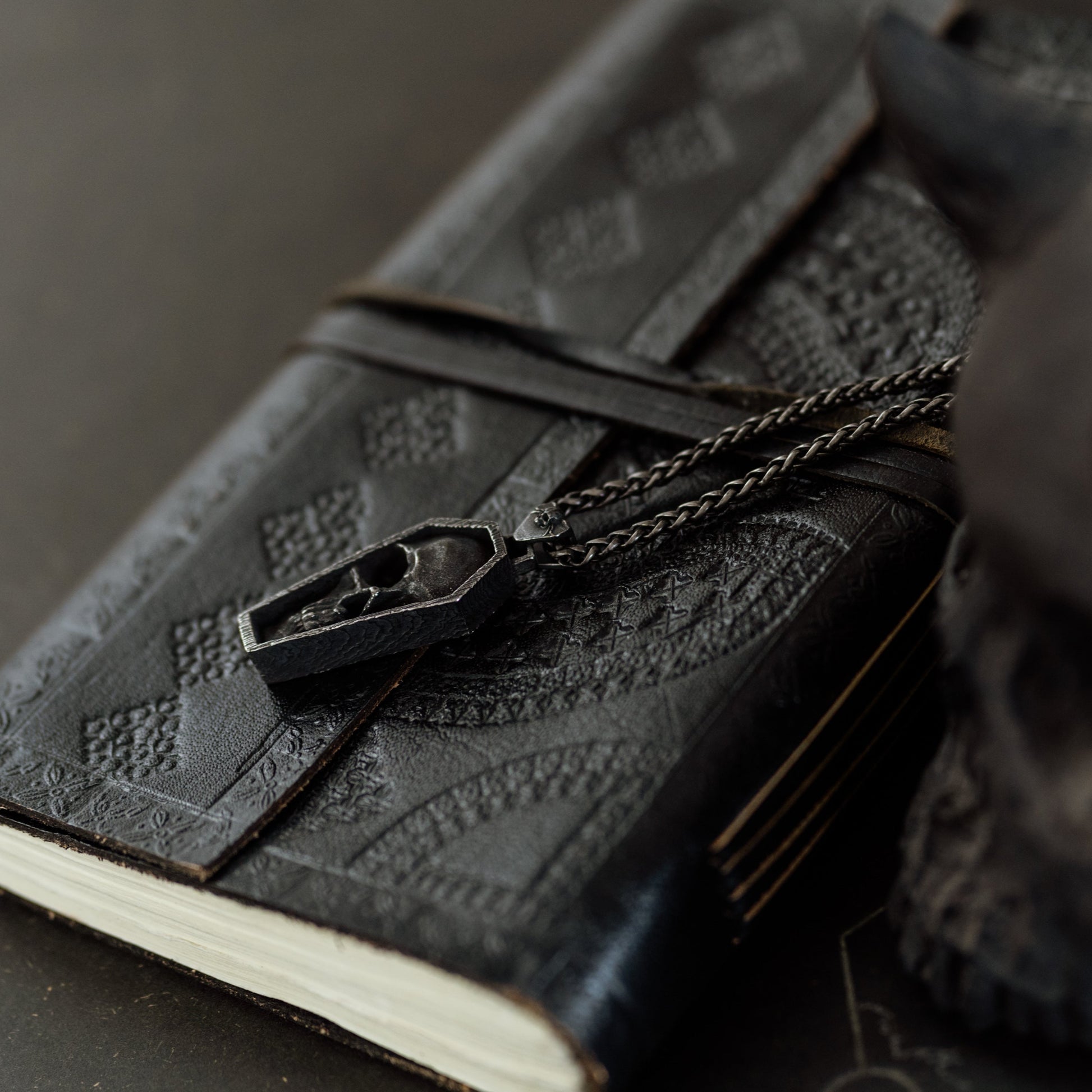 Black leather notebook with embossed design and chain, held by a dark-skinned hand on a dark surface.