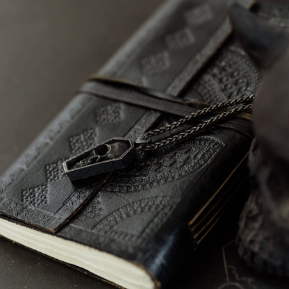 Black leather notebook with embossed design and chain, held by a dark-skinned hand on a dark surface.
